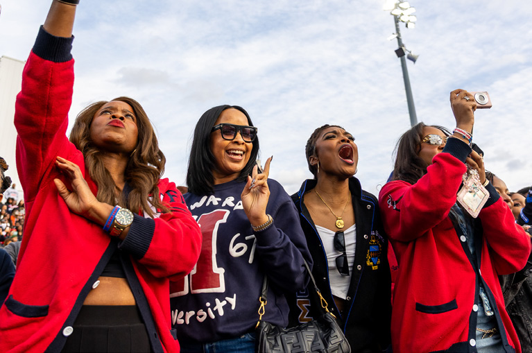 Alums wearing red and Howard University 1867 sweatshirts cheering at Homecoming on the Yard with a blue sky above.