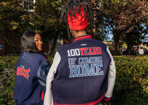 Students wearing Howard University jackets on The Yard and smiling. The jackets say,"100 Years of Coming Home" and "The Mecca"
