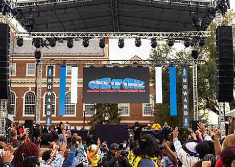 Crowd of students at Homecoming wearing colorful Howard University clothing and cheering for performers on the stage. A "One of One" Howard University Homecoming 2026 banner hangs on the stage with Founders Library in the background.